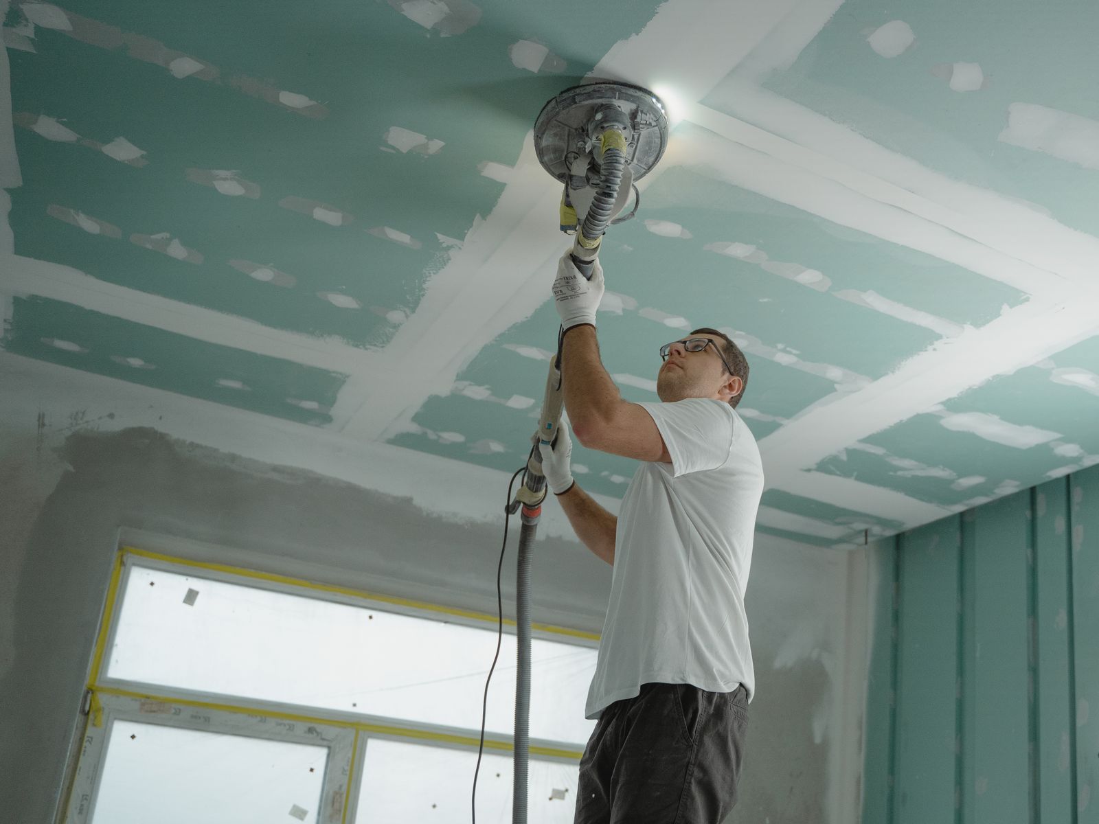 Worker using a round dust-extraction sander on a freshly taped drywall ceiling during a renovation