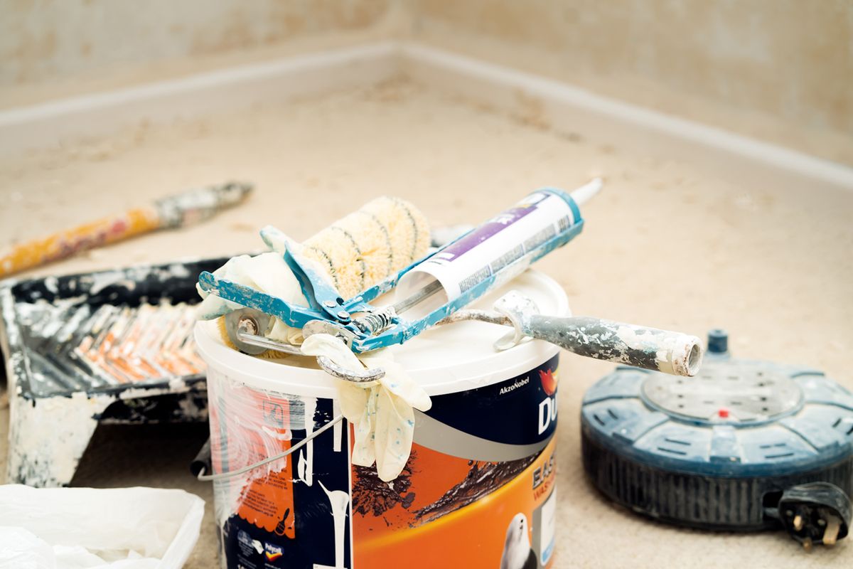 Paint bucket with a roller sleeve, caulk gun, and used painting tools resting on a protected floor